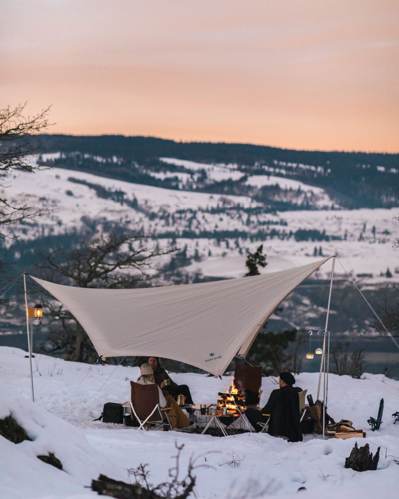 A group of people are gathered around a campfire beneath a tarp, enjoying the scenic winter landscape with snow-covered mountains in the background.